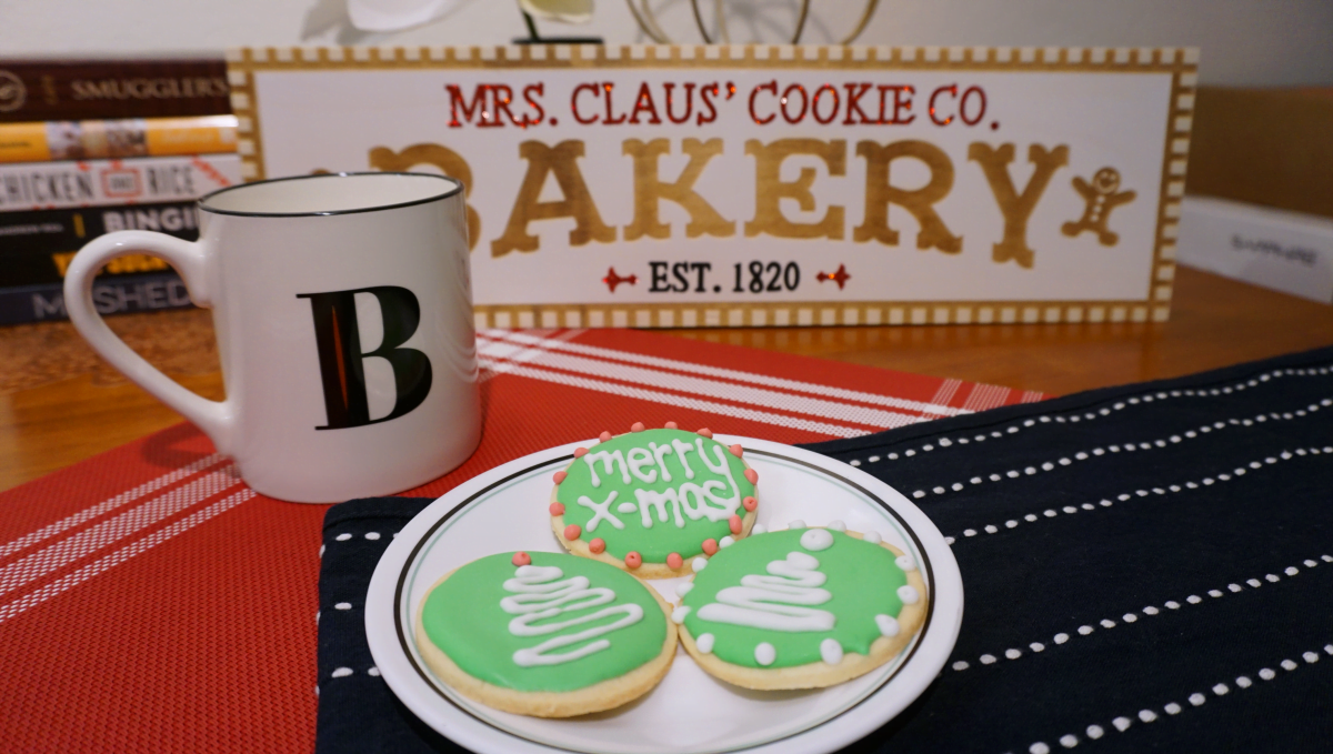 Plate of sugar cookies with green icing background and a white squiggle tree design on top. White mug with black letter B next to the plate of cookies. Sign behind the cookies and mug. Decorating Perfect Sugar Cookies | Sincerely Yasmin