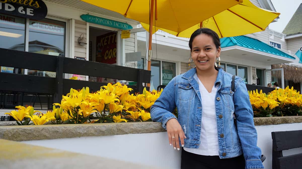 During a beach summer weekend visit to Cape May, Yasmin is standing in front of some yellow flowers while wearing a jean jacket and white shirt.