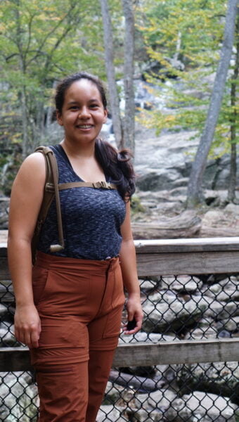 Yasmin standing in front of a waterfall while wearing a gray sports tank top and burnt orange hiking pants - REI Hiking Pants Review