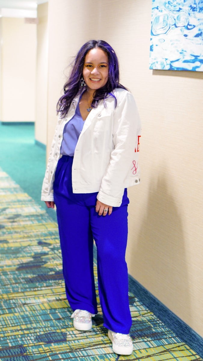 Yasmin is wearing a purple top with blue pants and a white jacket.